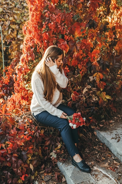 Jonge vrouw zit in het park in de herfst en houdt een boeket rode chrysantenbloemen vast. Ze stopt haar lange, steile haar achter haar oor.