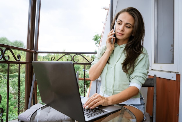 Jonge vrouw zit op een balkon aan een tafel met een telefoon achter een laptop