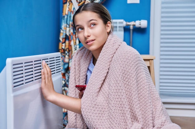 Jonge vrouw zit thuis bij een elektrische verwarmer tienermeisje verwarmt zich onder een deken verwarmingsseizoen energiecrisis besparing koude herfst winter seizoen concept