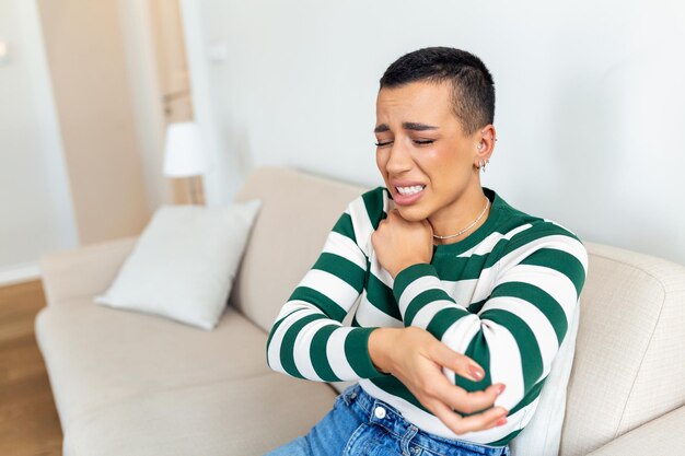 Jonge vrouw zit thuis op haar bank en raakt haar elleboog aan terwijl ze lijdt aan elleboogpijn