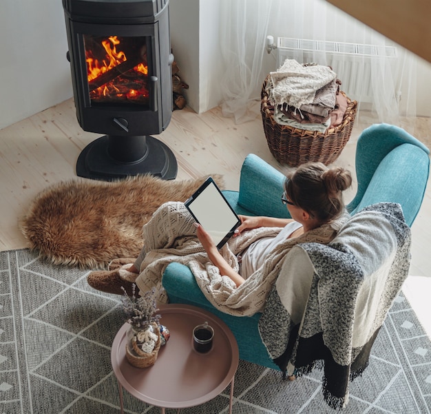 Jonge vrouw zitten in gezellige fauteuil, met warme deken, met behulp van tablet