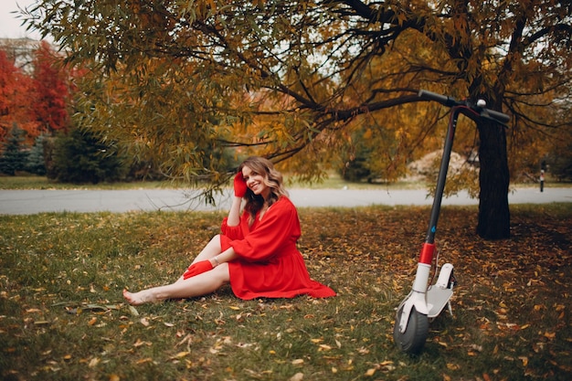 Jonge vrouw zitten met elektrische scooter in rode jurk in het herfst stadspark