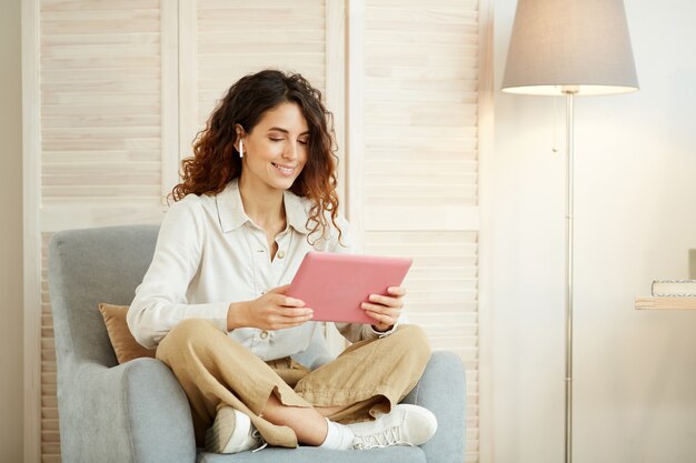 Jonge vrouw zittend in een stoel in de woonkamer en doet haar werk met behulp van tabletcomputer en internet