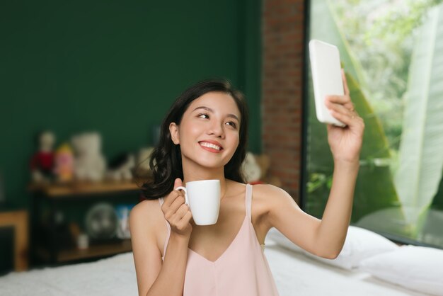 Jonge vrouw zittend op bed met kopje koffie selfie op mobiele telefoon