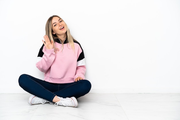 Jonge vrouw zittend op de vloer binnenshuis saluerend met de hand met gelukkige uitdrukking