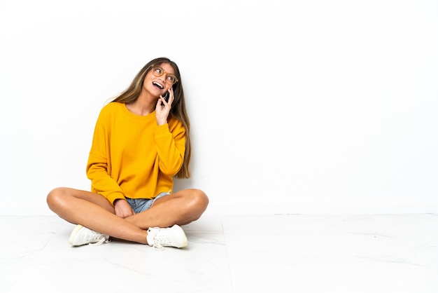 Jonge vrouw zittend op de vloer geïsoleerd op een witte achtergrond een gesprek te houden met de mobiele telefoon