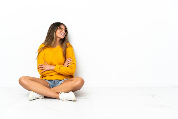 Jonge vrouw zittend op de vloer geïsoleerd op een witte achtergrond met gekruiste armen en gelukkig