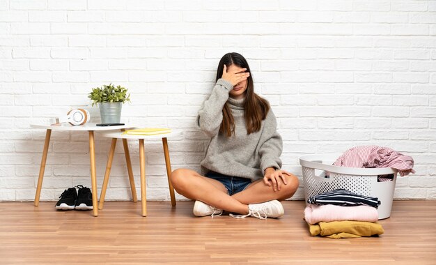 Jonge vrouw zittend op de vloer op binnenshuis met wasmand voor ogen door handen. Ik wil niets zien