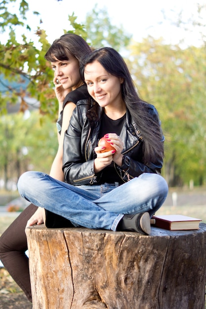 Jonge vrouw zittend op een afgezaagde boomstam buitenshuis eten van een appel terwijl haar vriend chats op haar mobiele telefoon
