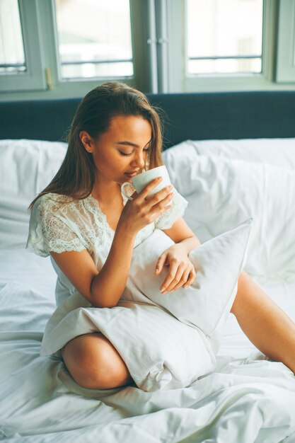 Jonge vrouw zittend op een bed en koffie drinken na het wakker worden