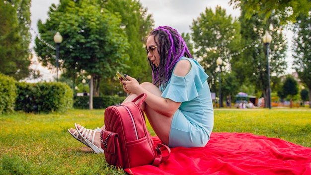 Jonge vrouw zittend op een deken in het park en het gebruik van een smartphone Gelukkige jonge brunette zittend op een groen gazon kijken naar inhoud op de telefoon op een zonnige dag