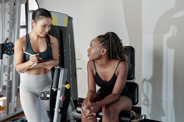 Jonge vrouw zittend op een gymmachine en luisterend naar de aanbevelingen van haar fitnesstrainer