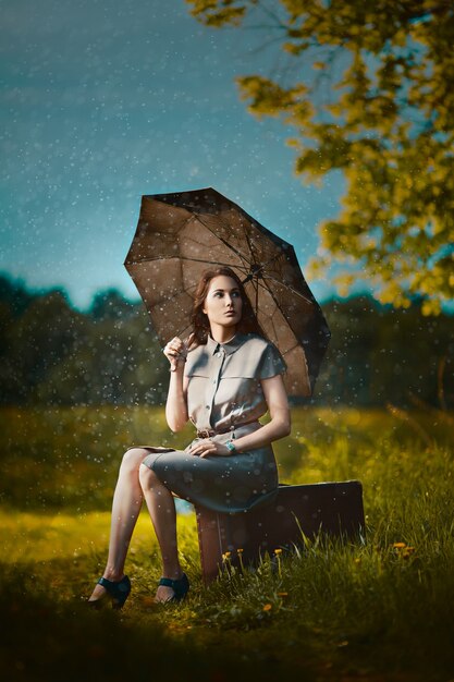 Jonge vrouw zittend op een koffer onder regen en wachten