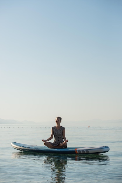 Jonge vrouw zittend op een paddleboard drijvend op een kalme ochtendzee en mediterend in een lotushouding
