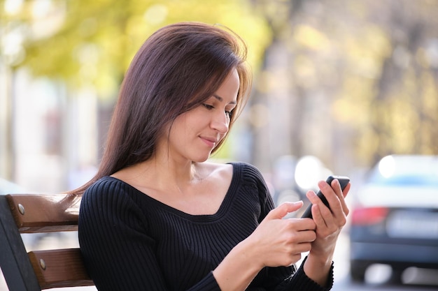 Jonge vrouw zittend op een straatbank die op een warme zomerdag buiten op haar mobiel bladert. Communicatie en mobiel verbindingsconcept.