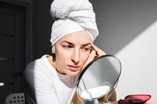 Jonge vrouw zonder make-up met een handdoek op haar hoofd plukt haar wenkbrauwen