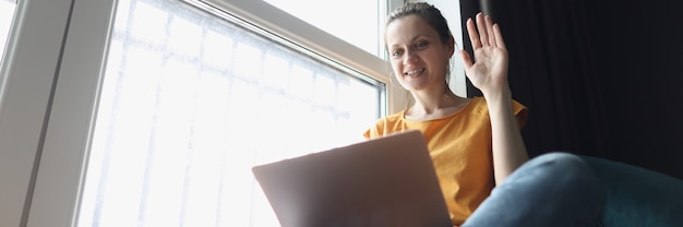 Foto jonge vrouw zwaait welkom op laptop terwijl ze op de vensterbank zit