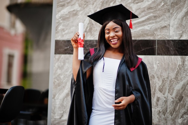 Jonge vrouwelijke Afro-Amerikaanse student met diploma poseert buitenshuis.