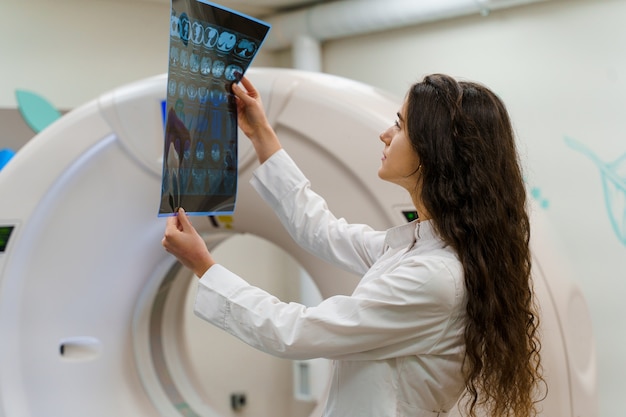 Jonge vrouwelijke arts in medische jurk kijkt naar resultaten naast CT-scanner