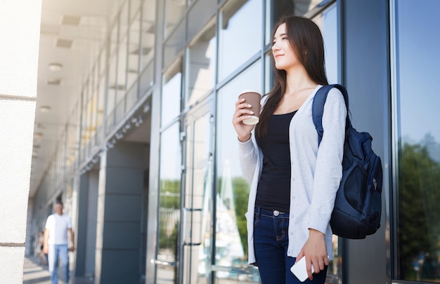 Jonge vrouwelijke aziatische student, met smartphone en rugzak, koffie drinken voor de les en glimlachen. Denkend aan de komende dag. Onderwijs, technologie, sociale media en gadgetverslavingsconcept