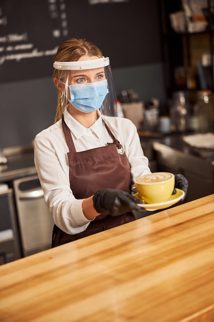 Jonge vrouwelijke barista in plastic schild en medisch masker met een kopje koffie boven de toog