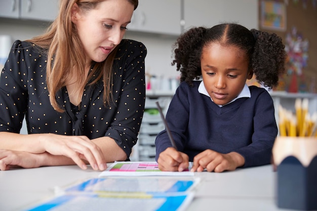 Jonge vrouwelijke basisschoolleraar werkt één op één met een schoolmeisje die haar aan een tafel in een klaslokaal van dichtbij ziet schrijven