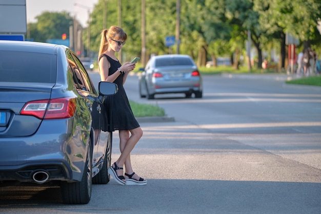 Jonge vrouwelijke chauffeur die in de zomer in de buurt van haar auto staat te praten op een mobiele telefoon op een straat in de stad