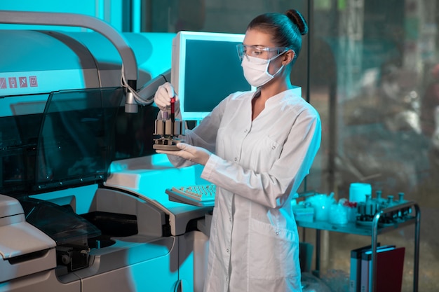 Jonge vrouwelijke chemicus die in een laboratorium werkt. Ze houdt in haar handen een set reageerbuisjes met bloed voor biochemische analyse in een modern apparaat.