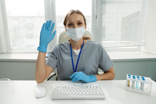 Jonge vrouwelijke clinicus of werknemer van medisch laboratorium in beschermende werkkleding die haar open handpalm naar u laat zien terwijl ze de elleboog op tafel houdt