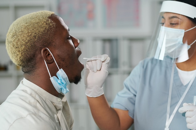 Jonge vrouwelijke clinicus van gemengd ras in beschermende werkkleding die voor de Afrikaanse man staat met open mond terwijl hij hem test op covid