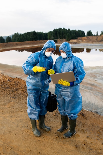 Jonge vrouwelijke ecologen in beschermende werkkleding die op vervuilde grond of klei op vuile rivier staan en aantekeningen maken in laptop