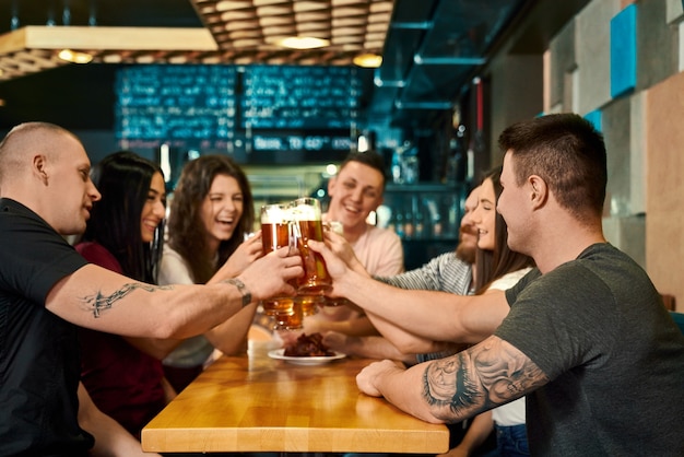 Jonge vrouwelijke en mannelijke vrienden die pinten met bier houden en in de pub roosteren. Gelukkig gezelschap aan tafel zitten, elkaar aankijken, lachen en praten in café. Concept van brouwerij en plezier.