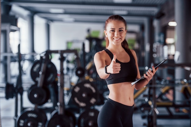 Jonge vrouwelijke fitness personal trainer met Kladblok staande in de sportschool met duim omhoog.