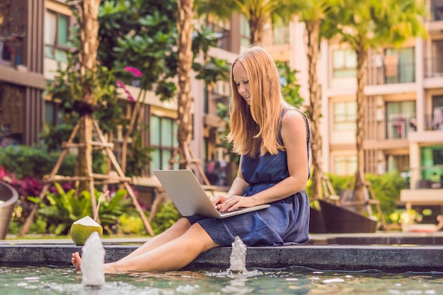 Jonge vrouwelijke freelancer zit bij het zwembad met haar laptop in het hotel en bladert op haar smartphone. Druk op vakantie. Verre werk concept. Kopieer ruimte voor uw tekst