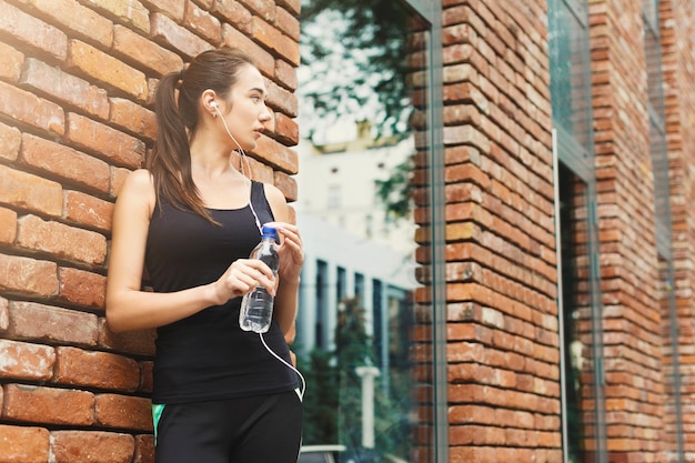 Jonge vrouwelijke hardloper heeft pauze, drinkt water tijdens het joggen in het stadscentrum, bakstenen muurachtergrond, kopieer ruimte