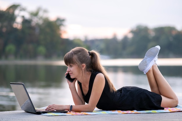 Jonge vrouwelijke kantoormedewerker die achter een laptop werkt terwijl hij op een mobiele telefoon praat terwijl hij in het zomerpark in de buitenlucht ligt. werkconcept op afstand