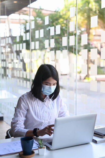 Jonge vrouwelijke kantoormedewerker met beschermend masker werkt of controleert informatie van de website op haar laptop op kantoor.