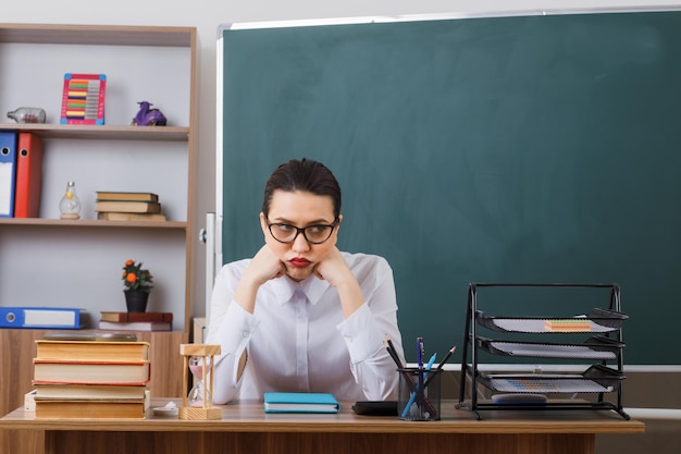 Jonge vrouwelijke leraar die een bril draagt en uitleg geeft over de les die er moe en overwerkt uitziet aan de schoolbank voor het bord in de klas