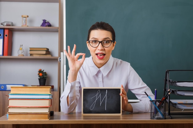 Jonge vrouwelijke leraar die een bril draagt met een klein schoolbord dat aan de schoolbank zit voor het bord in de klas en naar de camera kijkt met een goed teken dat lacht