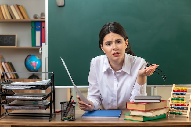 jonge vrouwelijke leraar die papier vasthoudt met een bril die aan tafel zit met schoolhulpmiddelen in de klas