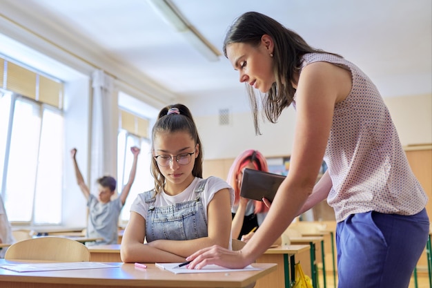 Jonge vrouwelijke leraar leert les in de klas van tienerkinderen leraar staat in de buurt van schoolbank met meisje student controleert kennis Onderwijs school college onderwijsconcept