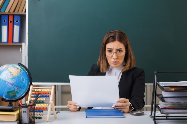 Jonge vrouwelijke leraar met een bril die aan de schoolbank zit met globe en boeken voor het bord in de klas met een wit leeg vel papier dat er ontevreden uitziet