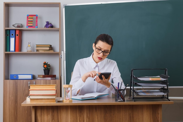 Jonge vrouwelijke leraar met een bril die aan de schoolbank zit voor het bord in de klas met behulp van een rekenmachine met een serieus gezicht