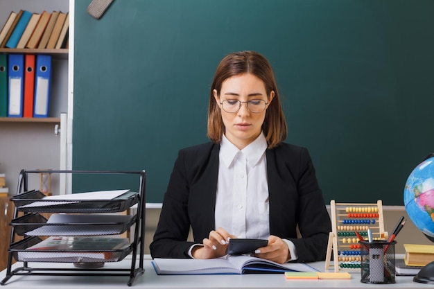 Jonge vrouwelijke leraar met een bril die aan de schoolbank zit voor het schoolbord in de klas met behulp van een rekenmachine die zich voorbereidt op een les die er zelfverzekerd uitziet