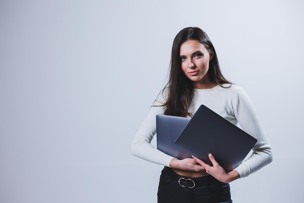 Jonge vrouwelijke manager in een wit overhemd houdt een draagbare laptop in haar handen terwijl ze op een grijze achtergrond staat