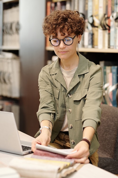 Jonge vrouwelijke manager van studio voor interieurontwerp die laptop gebruikt terwijl ze door nieuwe voorbeelden van stoffen kijkt voor het maken van meubels