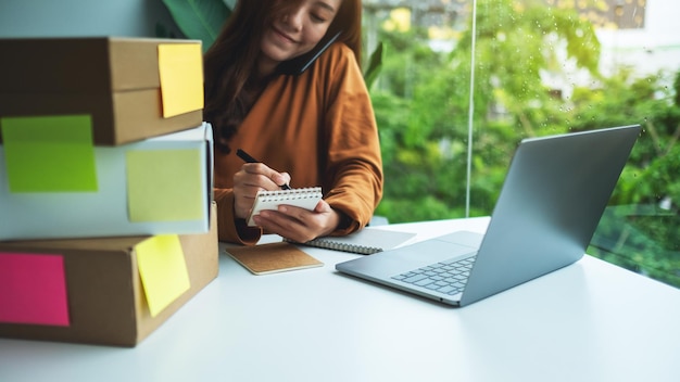 Jonge vrouwelijke ondernemer die bestellingen van de klant aan de telefoon bevestigt en noteert voor online winkel- en bezorgconcept