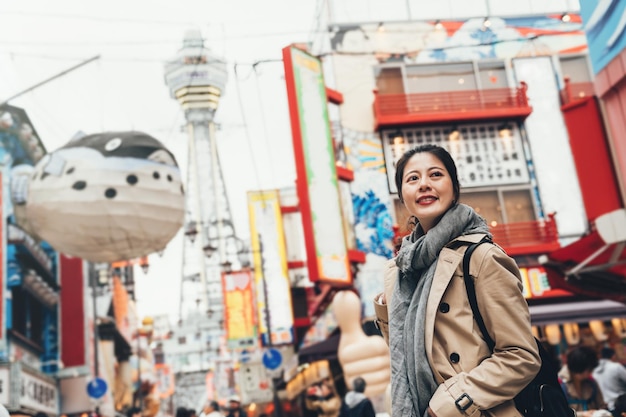 jonge vrouwelijke reiziger die op zonnige dag vrolijk op de krioelende straat in tsutenkaku staat en een sjaal draagt. enorme kogelvisballon die in de lucht vliegt. meisje glimlachend rondkijken op de weg.