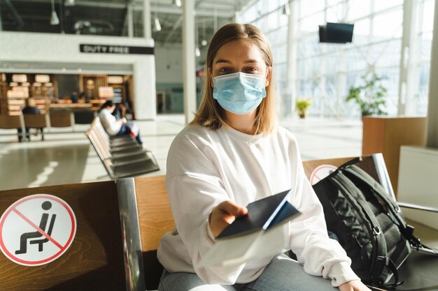 Foto jonge vrouwelijke reiziger in medisch masker zit in de wachtkamer en houdt paspoort en ticket uit