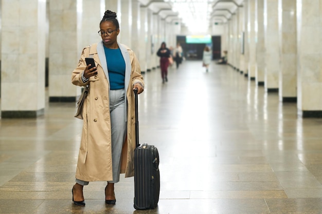 Jonge vrouwelijke reiziger met koffer die sms't in smartphone terwijl hij voor de camera staat